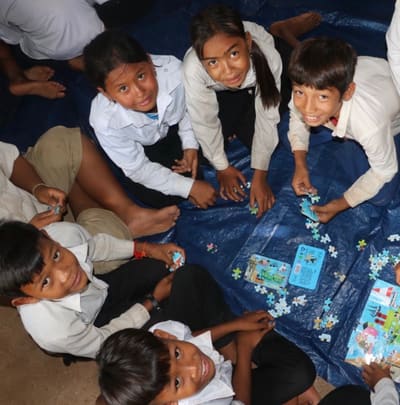 Schoolchildren sit together on a mat solving jigsaw puzzles, looking up and smiling