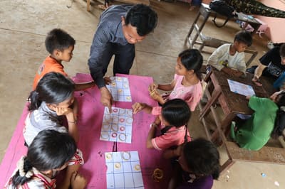 Children gather around a teacher for a counting game at a low pink table
