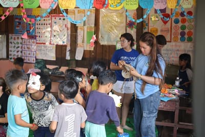 Children in a brightly decorated classroom take part in a crafts activity with a visiting helper