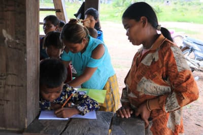 A woman helps a young boy practise writing at a village learning session